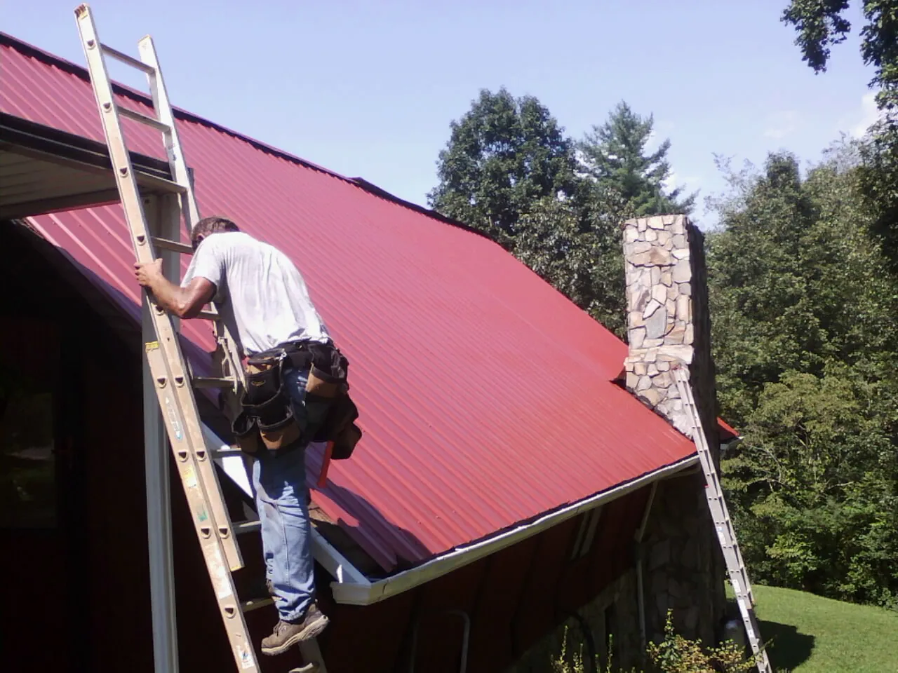 Roofer on a ladder beside a red metal roof and stone chimney — Soffit & Fascia Repair by Roofing Brothers in Taylor